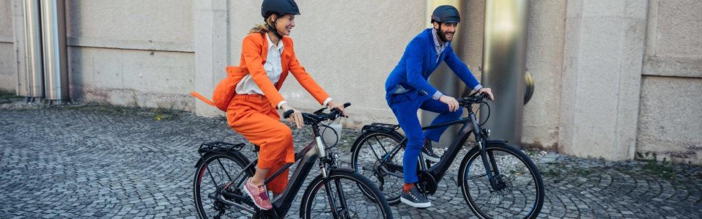 Das Bild zeigt zwei Personen, die auf modernen E-Bikes durch eine Stadtlandschaft fahren. Die Frau in einem orangef