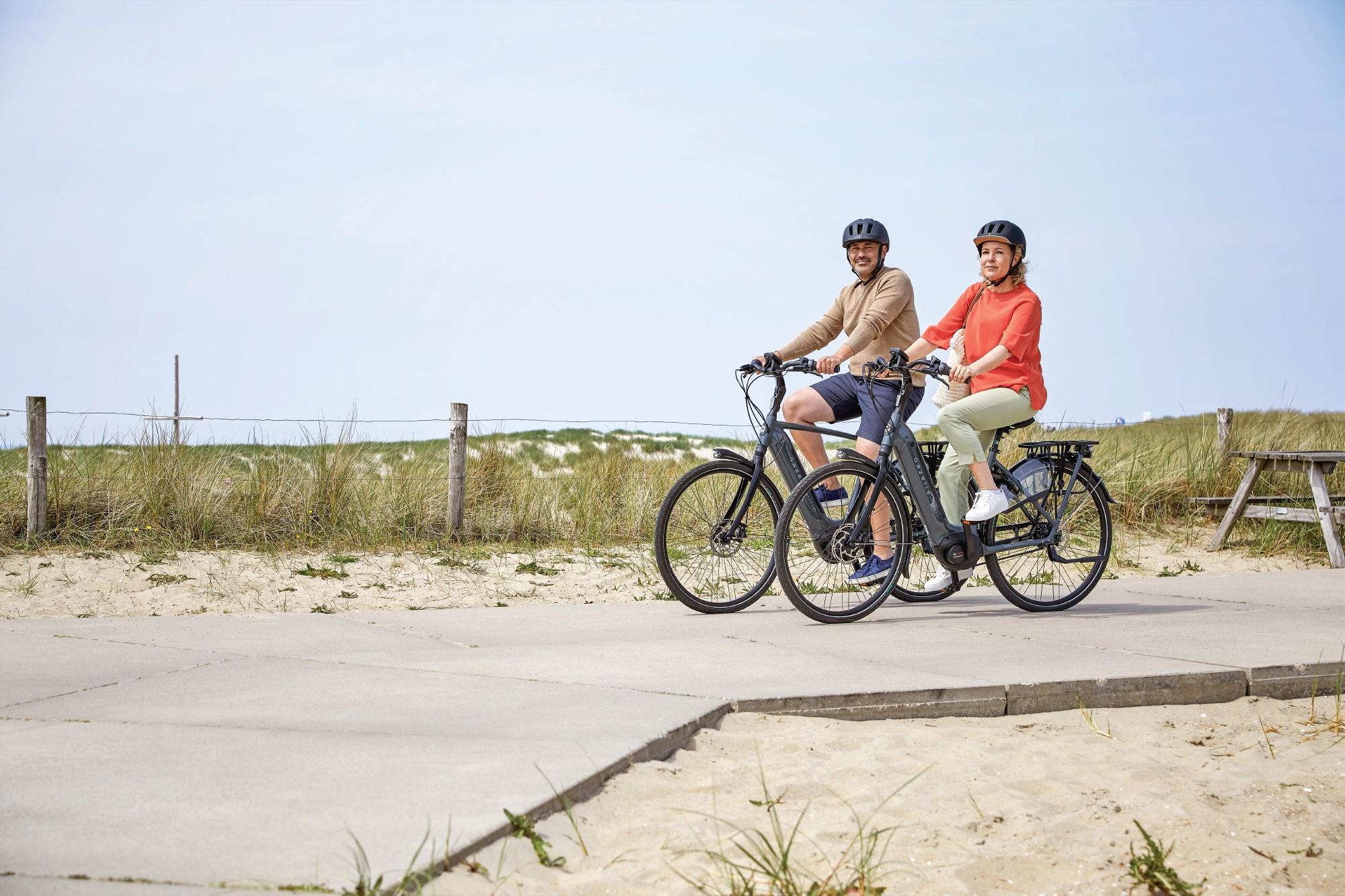 Das Bild zeigt zwei E-Bikes der Marke Gazelle, die in einer malerischen Strandlandschaft positioniert sind.