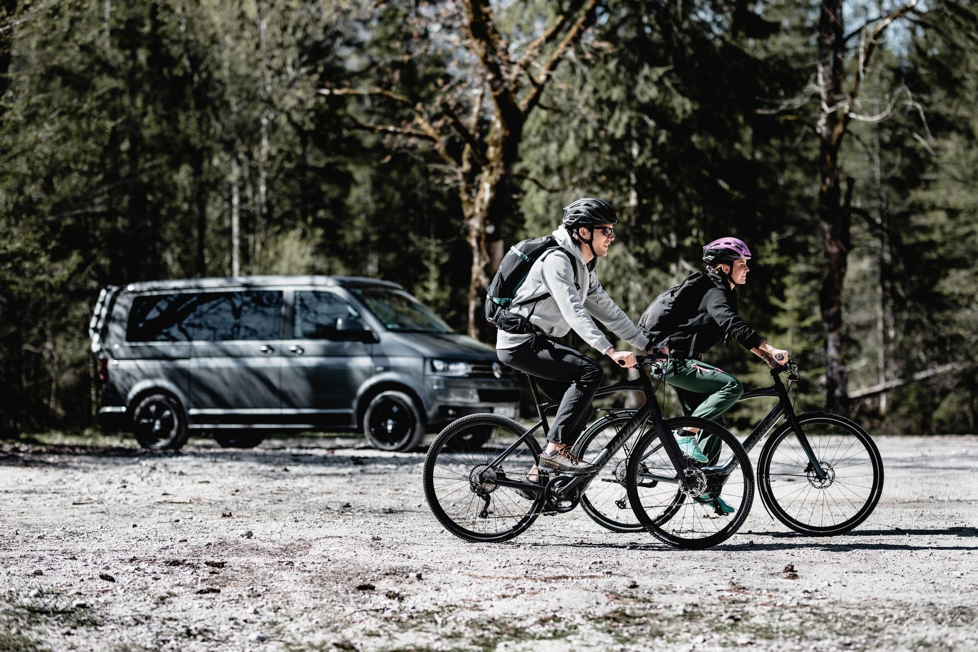 Zwei Personen fahren auf e-Bikes durch die Natur, im Hintergrund sieht man einen Transporter