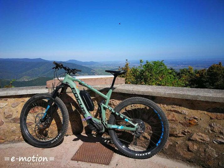 Ein E-Mountainbike der Marke Focus, Modell Jam, steht an einer Steinmauer mit Blick auf eine bergige Landschaft im