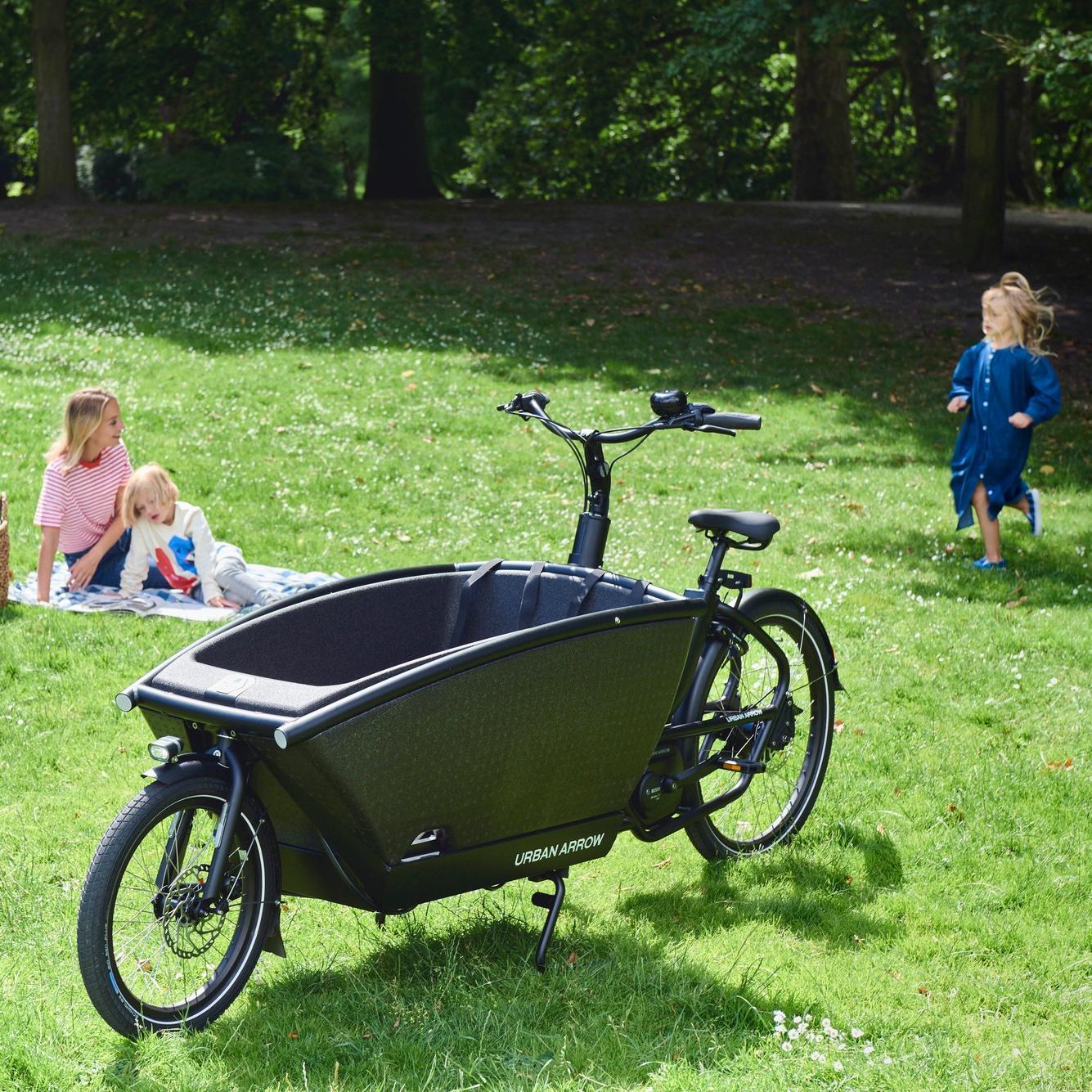 Ein Urban Arrow Family, daneben sitzt eine Mutter mit ihren beiden spielenden Kindern