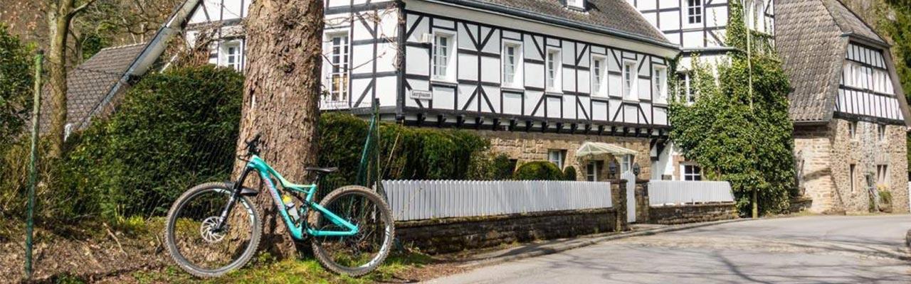 Ein Mountainbike lehnt an einem Baum vor einem idyllischen Fachwerkhaus in Wuppertal. Perfekt für Ausflüge durch di