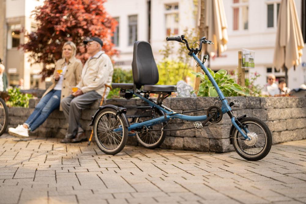 Ein blaues Pfautec-Dreirad steht im Vordergrund, zwei Personen entspannen sich im Hintergrund. Ideal für Mobilität