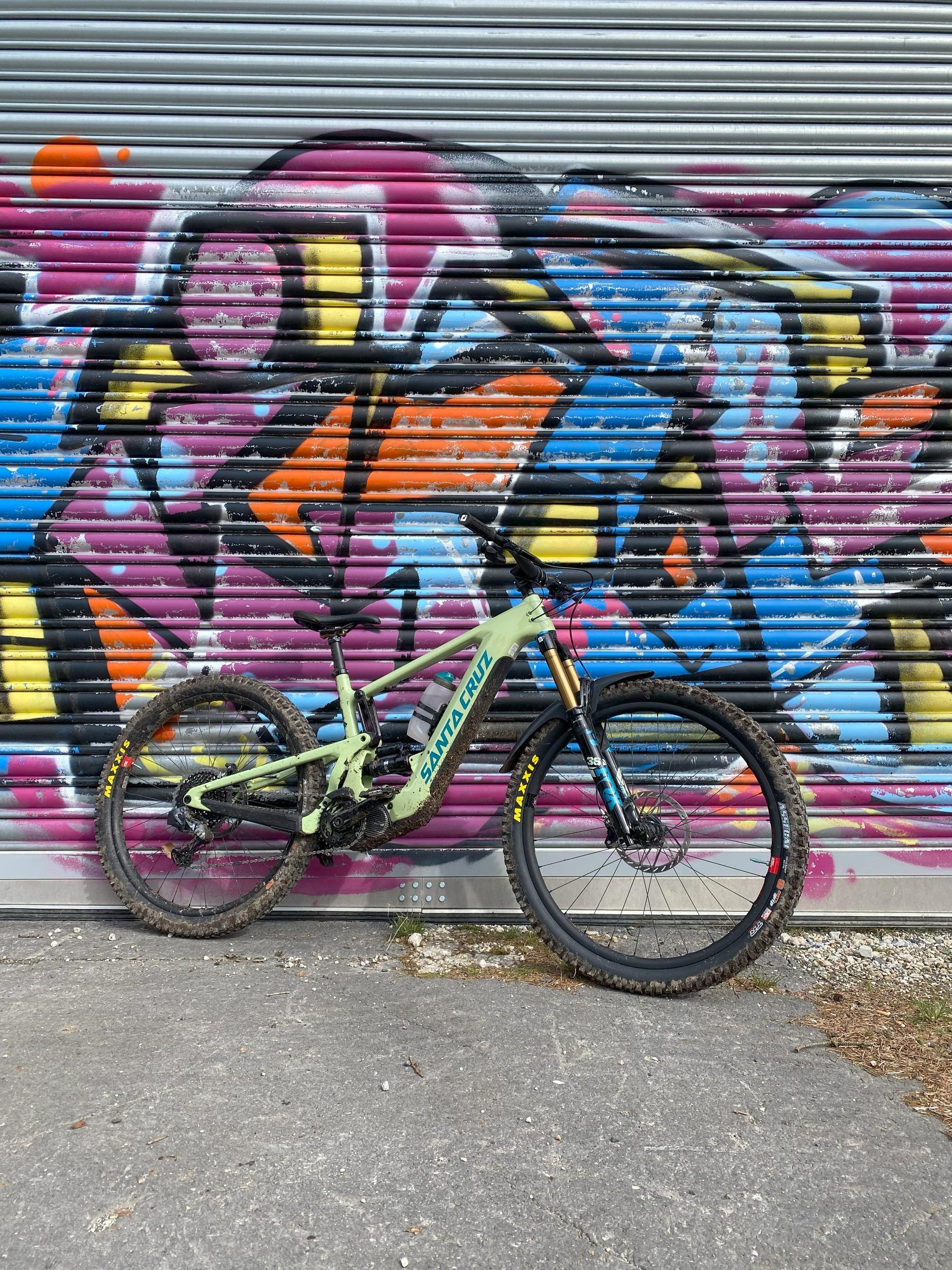 Das Bild zeigt ein E-Mountainbike der Marke Santa Cruz, das vor einer bunten Graffiti-Wand abgestellt ist.