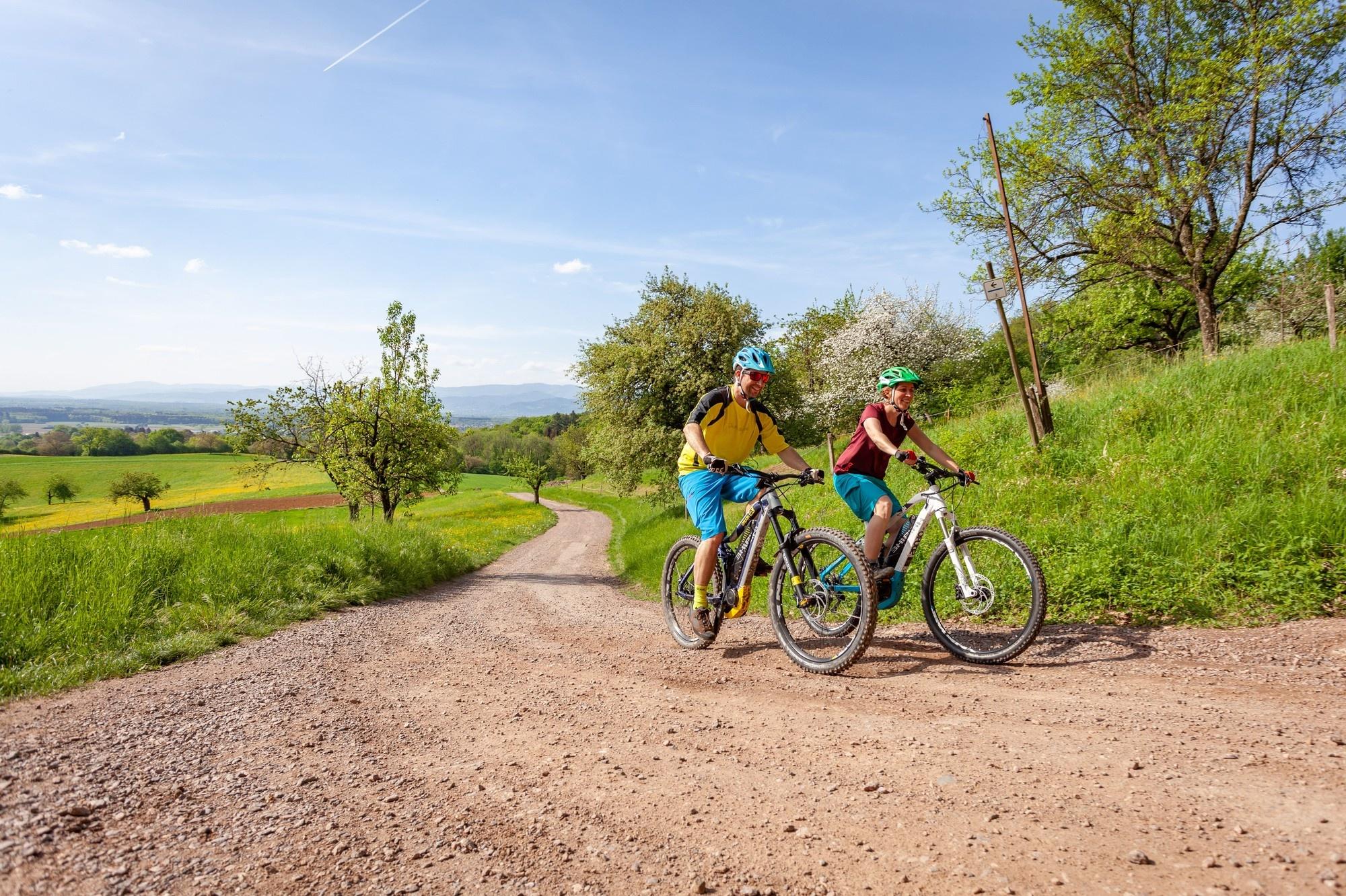 Das Bild zeigt zwei Radfahrer, die auf einem Schotterweg in einer grünen Landschaft fahren. Sie nutzen E-Mountainbi