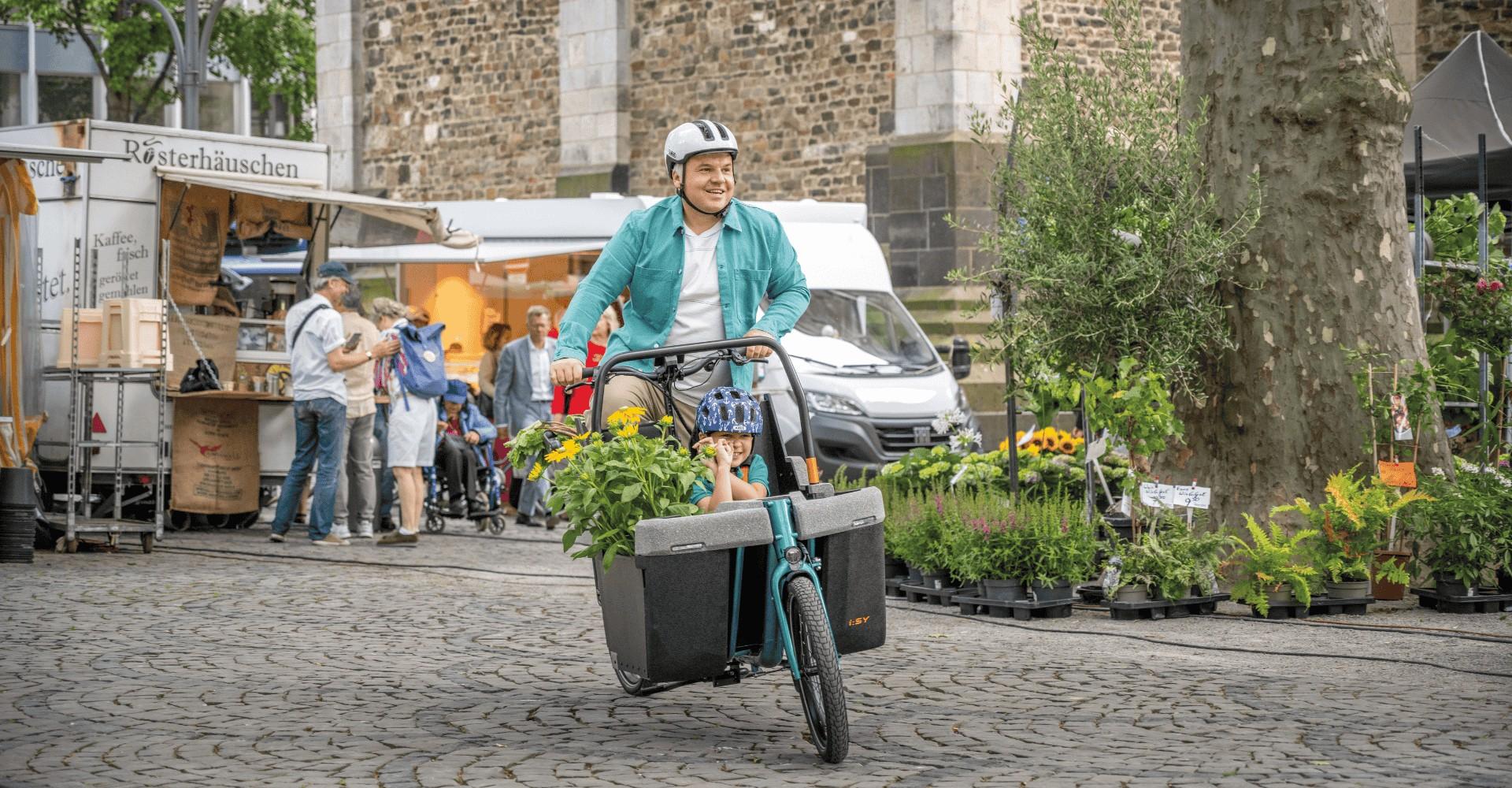 Ein Mann fährt mit dem i:SY Cargo P12 Maxi - in der Transportbox befindet sich ein Kind und Blumen