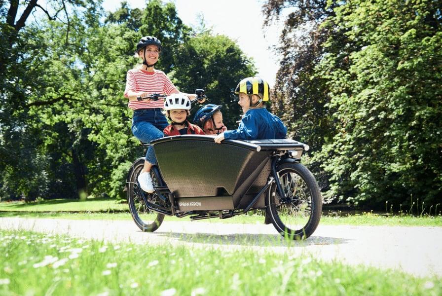 Eine Frau fährt mit drei Kindern auf einem Urban Arrow Family Lasten-E-Bike auf einem sonnigen Parkweg. Alle tragen