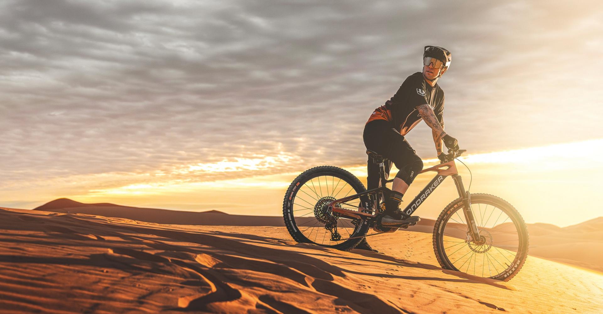 Ein Mann steht mit seinem e-Mountainbike auf einer Sanddüne in einer Wüste bei Sonnenuntergang