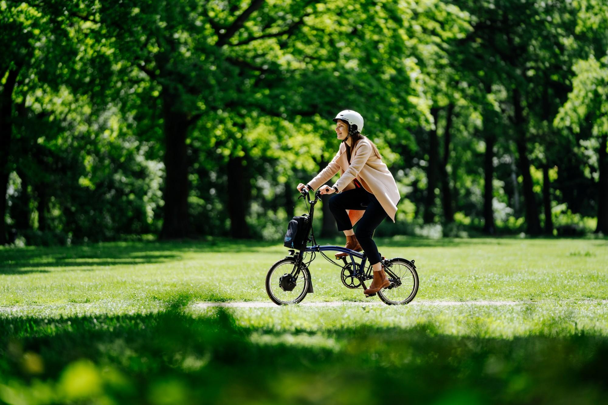 Das Bild zeigt eine Frau, die auf einem Brompton-Faltenfahrrad fährt, umgeben von einer grünen Parklandschaft.
