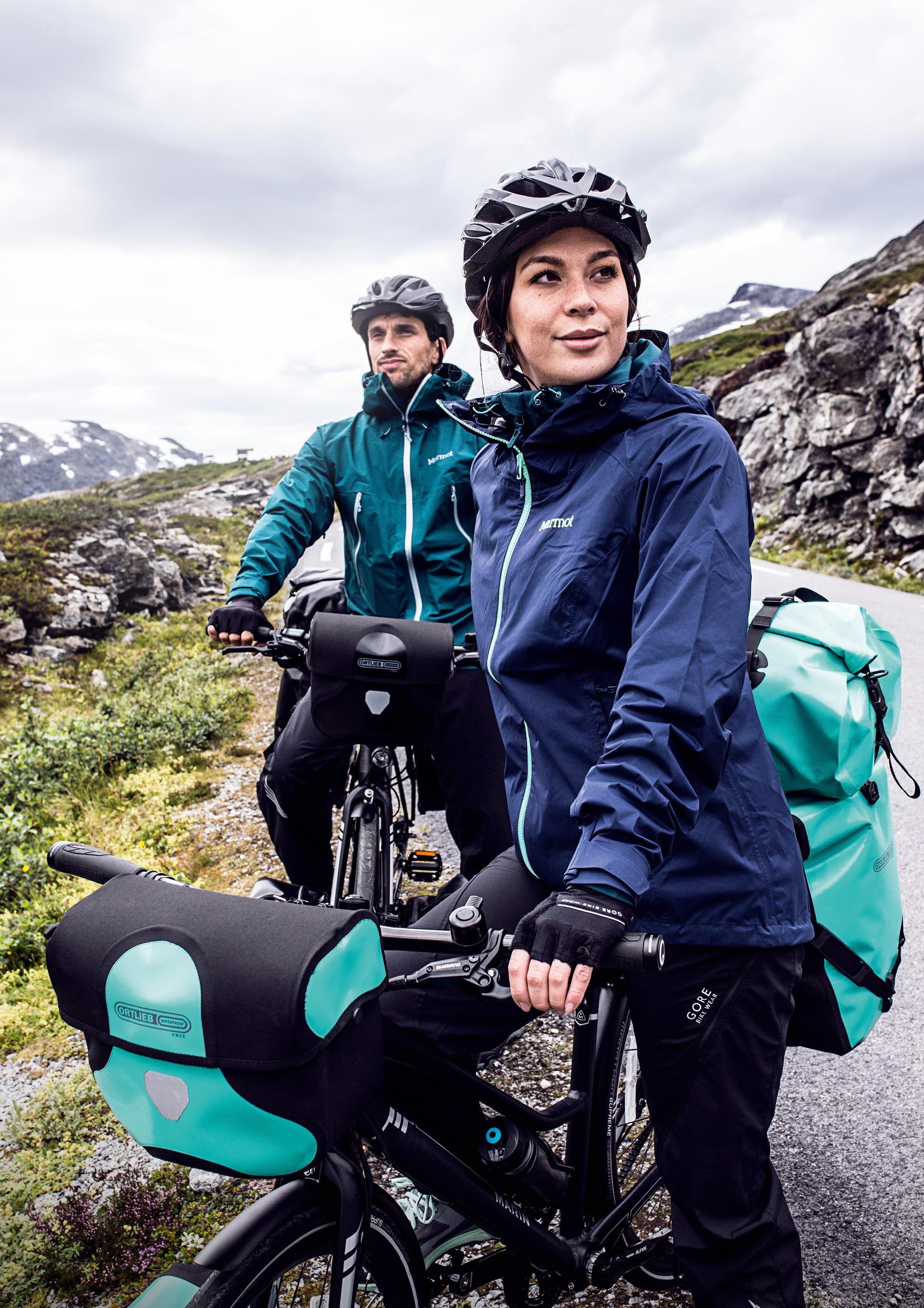 Frau steht mit ihrem e-Bike mit Ortlieb-Lenkertasche auf einer Straße in den Bergen.