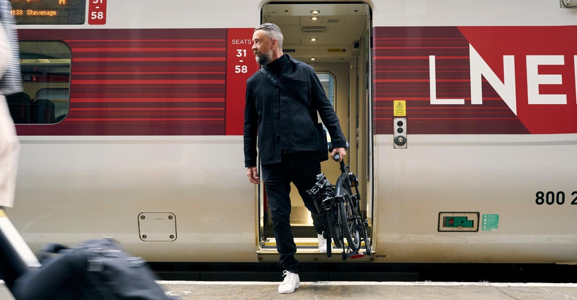 Das Bild zeigt einen Mann, der mit einer zusammengefalteten P Line von Brompton aus einem Zug steigt. Die P Line is