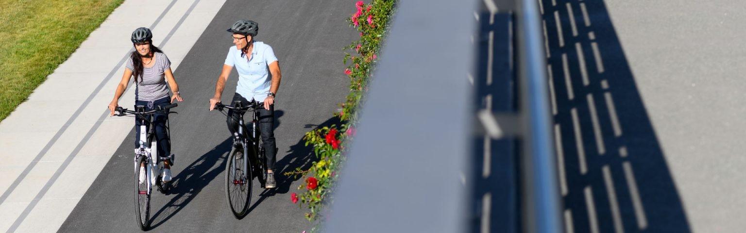 Das Bild zeigt ein Paar, das gemeinsam auf Winora Sinus Tria E-Bikes fährt. Beide Radfahrer tragen Helme zur Sicher