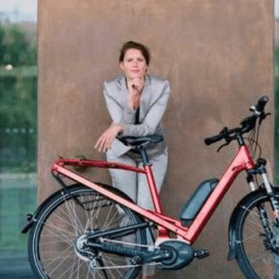 Frau in Business-Kleidung lehnt an rotem E-Bike vor Bürogebäude