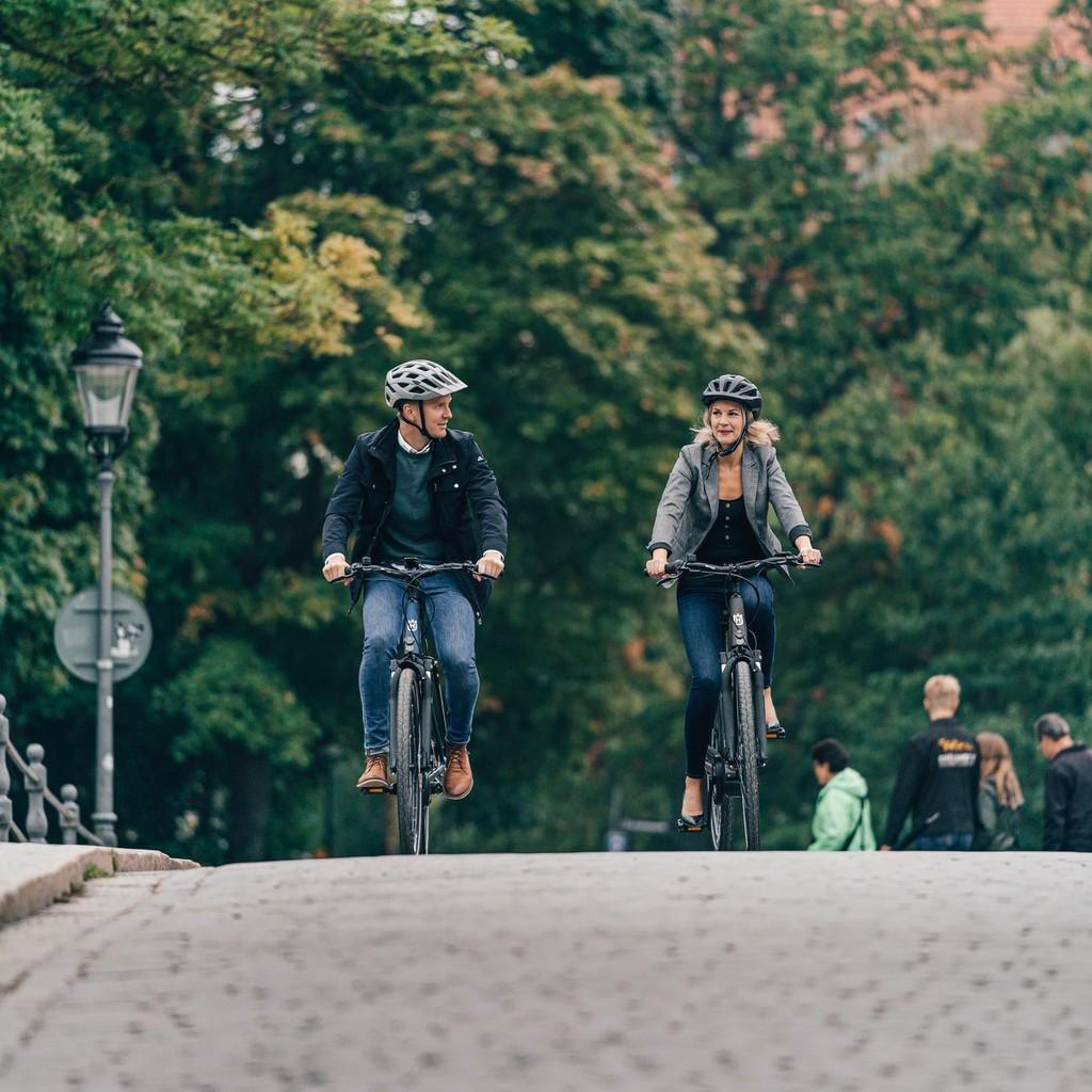 Zwei Personen fahren Eco City E-Bikes auf einer Brücke, umgeben von städtischem Grün.