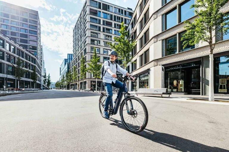 Das Bild zeigt einen Radfahrer auf einem modernen E-Bike, der durch eine weitläufige, urbane Straße fährt. Im Hinte