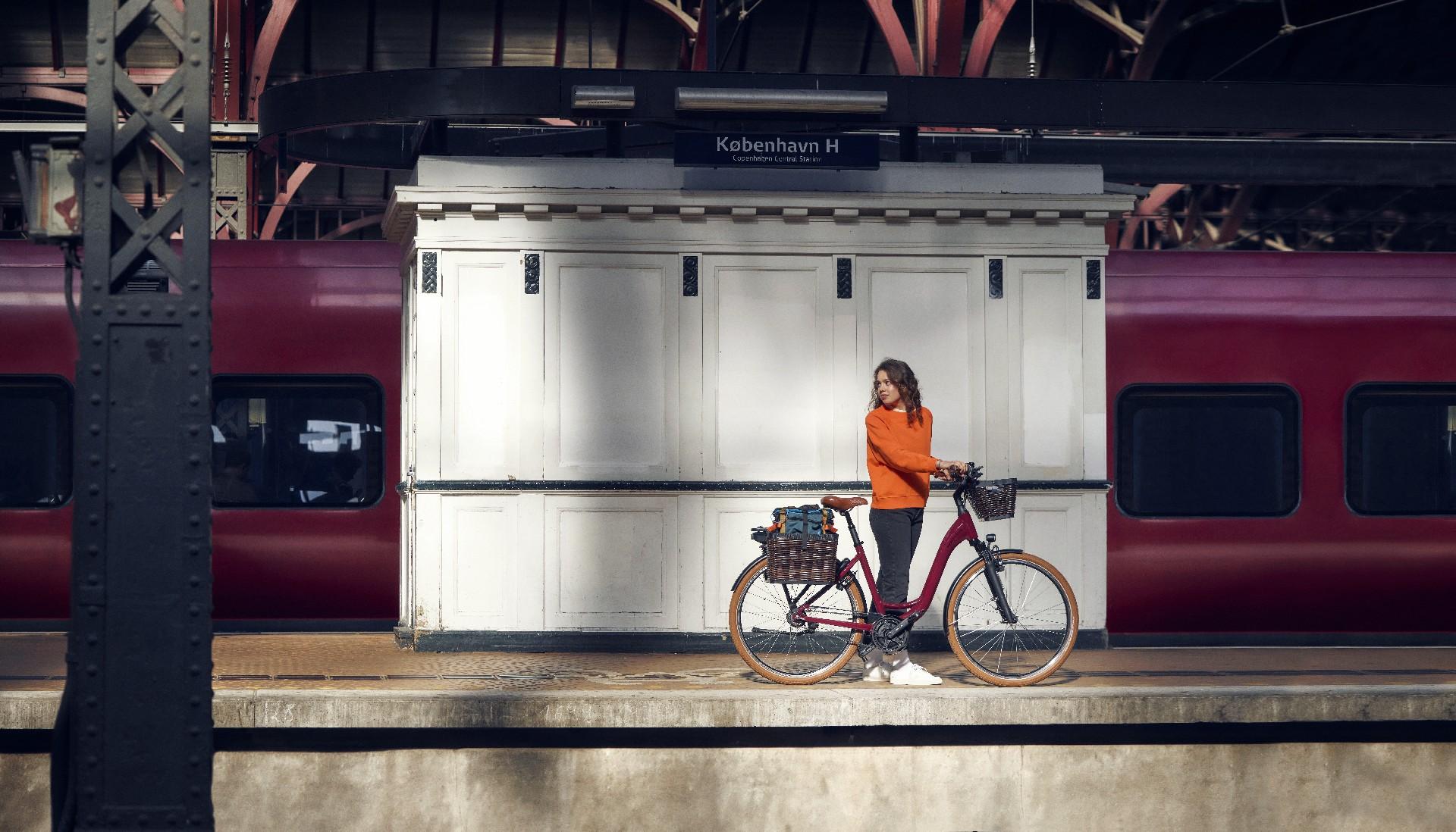 Riese & Müller Swing4, rotes Cargo-Bike mit Frontkorb am Bahnsteig. Daneben Person in Orange, hinten roter Zug.