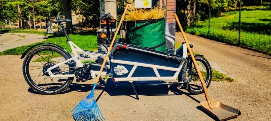 Weißes E-Bike Lastenrad vor einer Hecke mit Besen und Schaufel