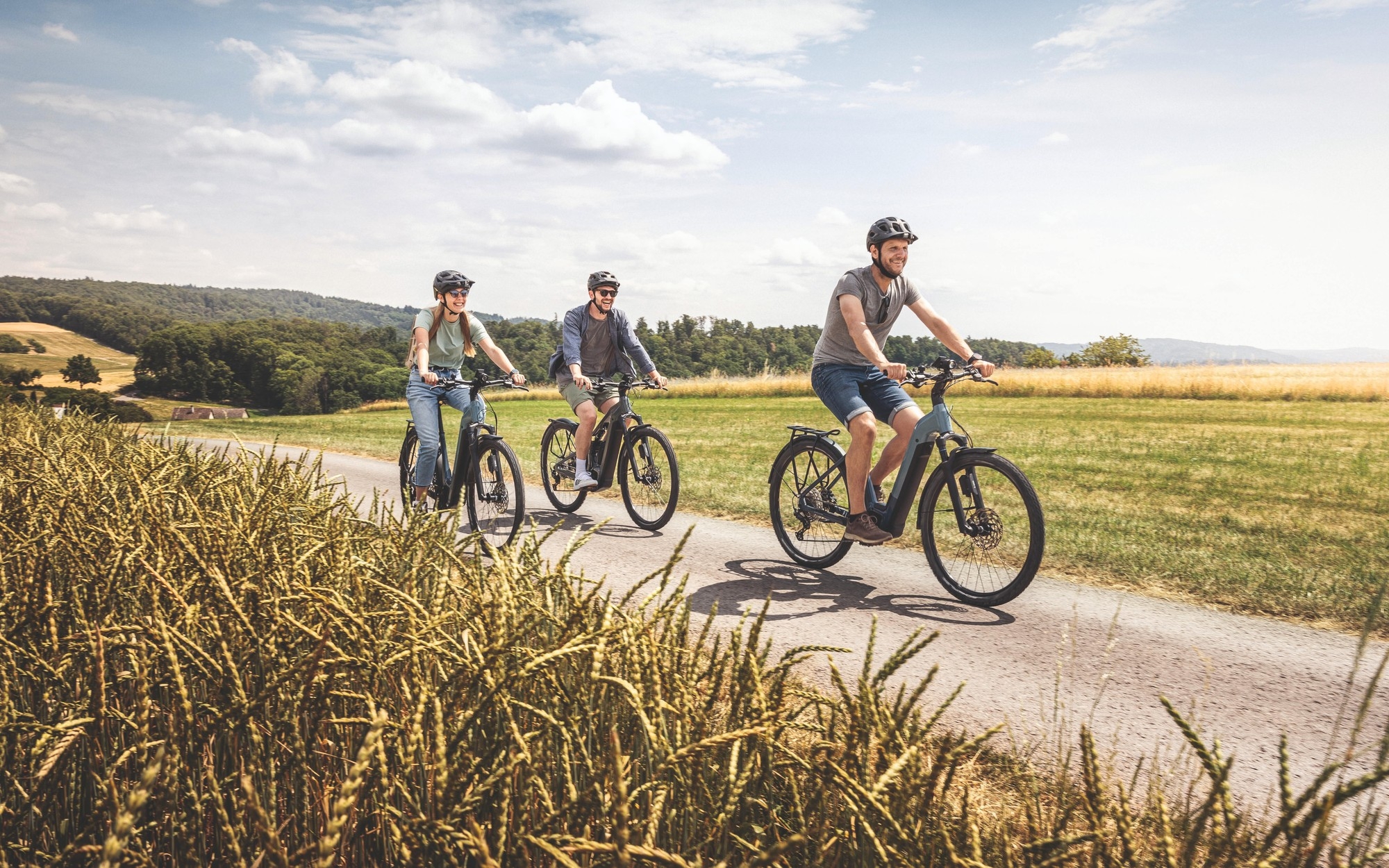 Das Bild zeigt drei E-Bike-Fahrer, die entspannt auf einem Asphaltweg durch eine malerische Landschaft fahren.