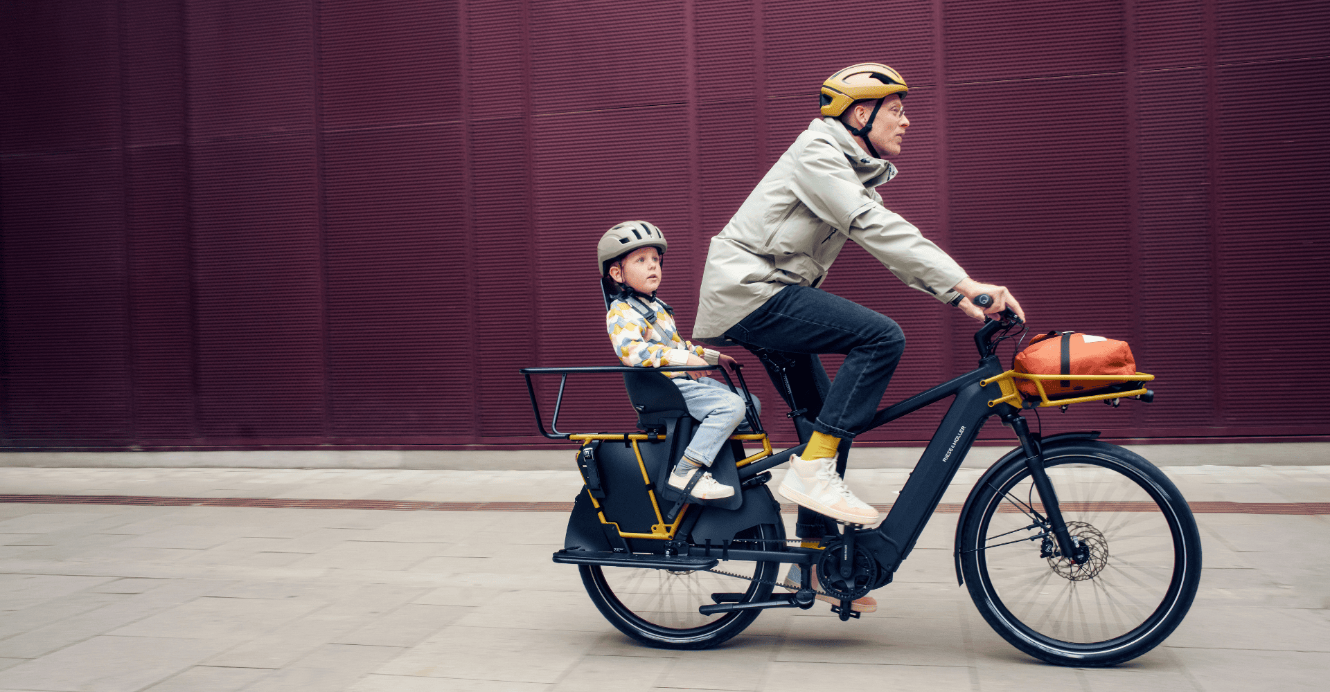 Ein Erwachsener fährt ein Lasten-E-Bike mit Kindersitz, in dem ein Kind mit Helm sitzt, vor roter Wand.