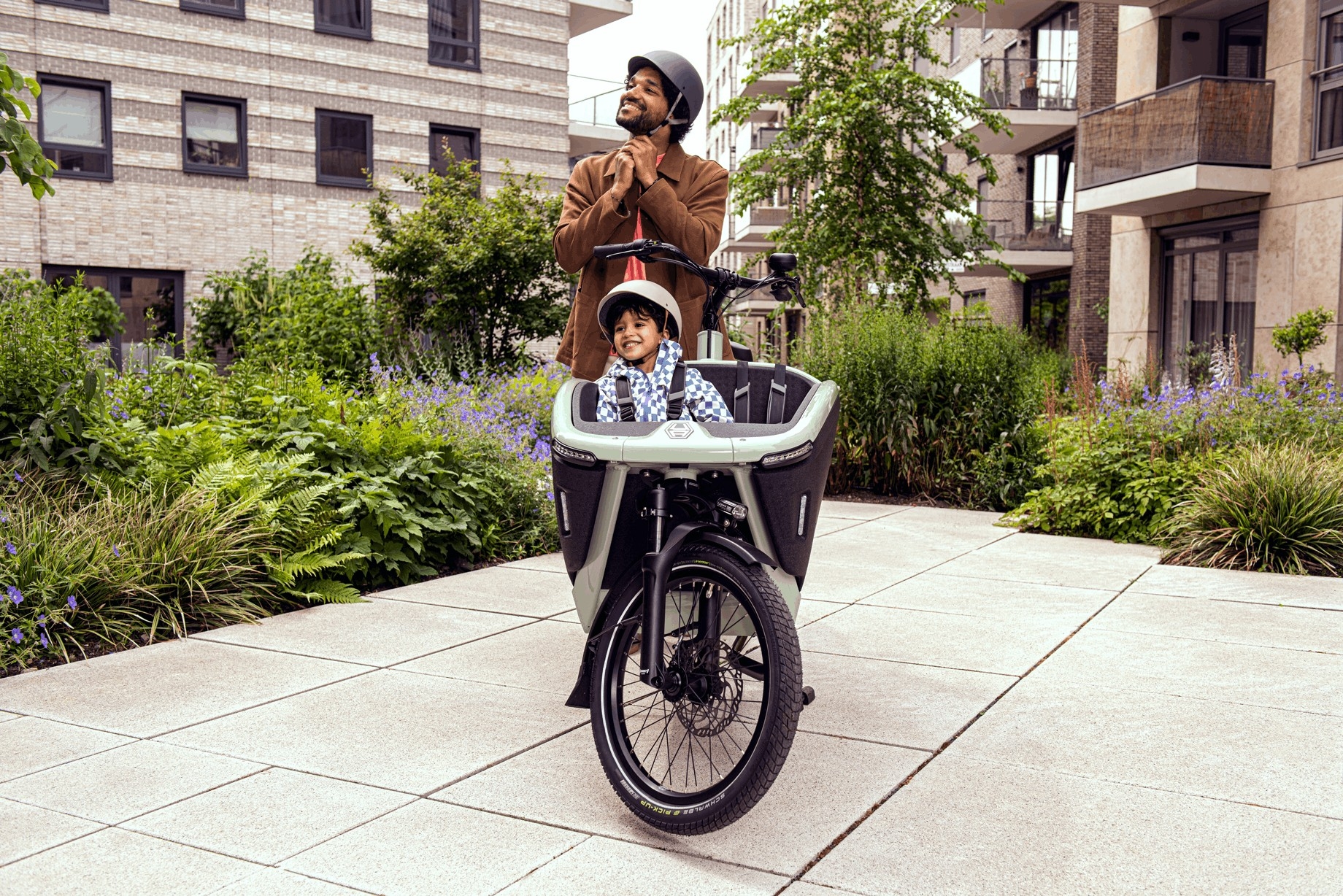 Vater mit Kind in einem Lastenrad auf einem Weg zwischen modernen Wohngebäuden.