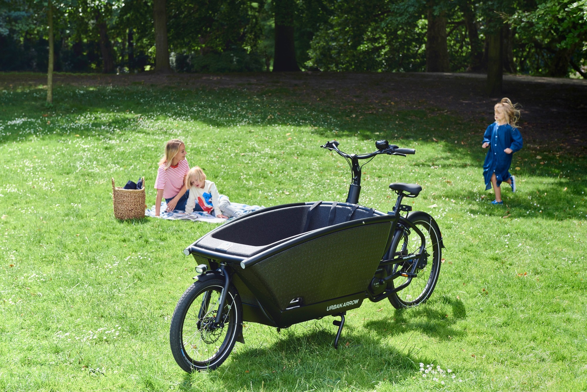 Ein Urban Arrow Family, daneben sitzt eine Mutter mit ihren beiden spielenden Kindern