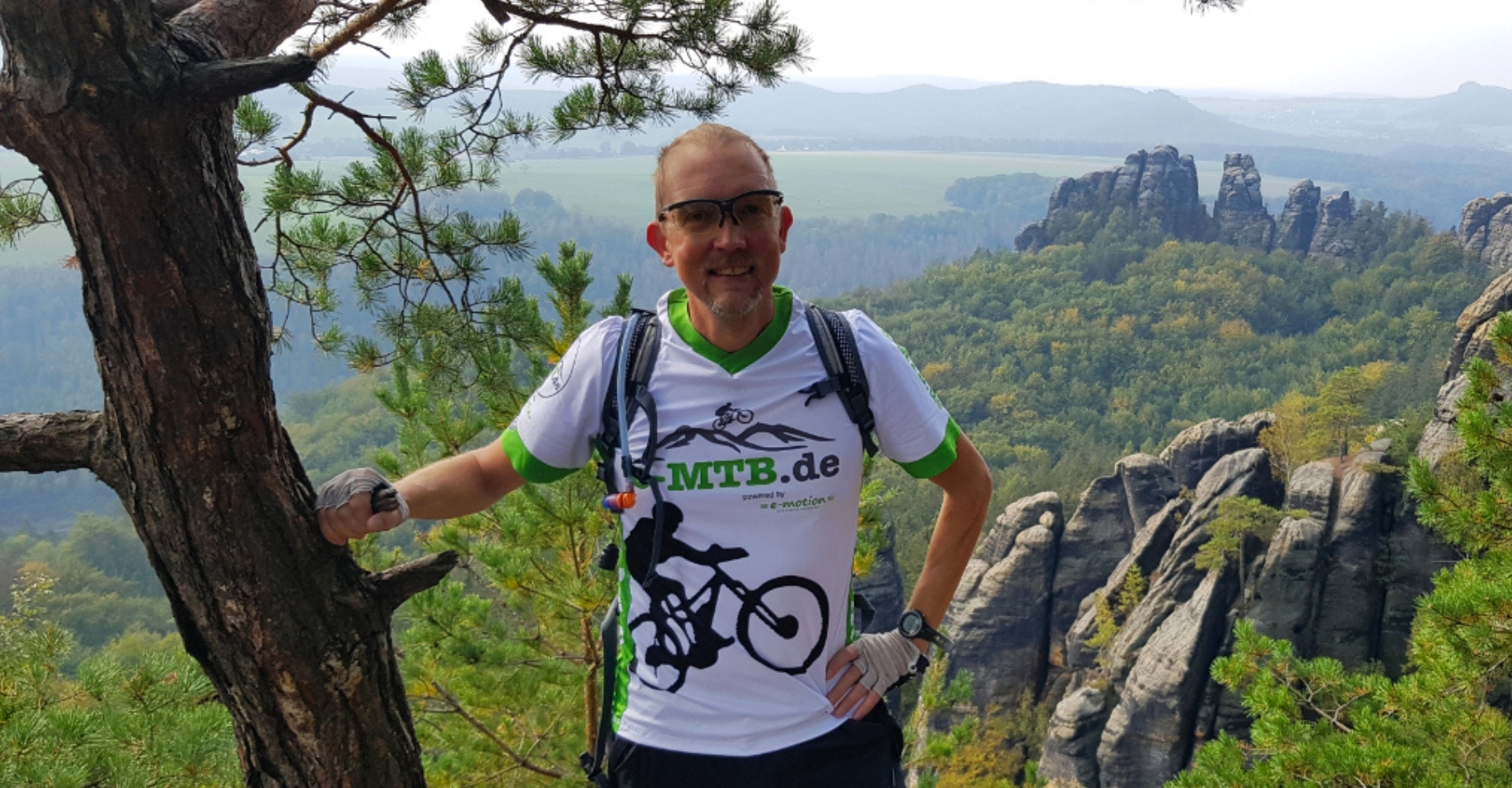 Der Kopf hinter Belchenradler, Christoph Steier, steht bei einer Radtour an einem Baum gelehnt mit Bergen dahinter