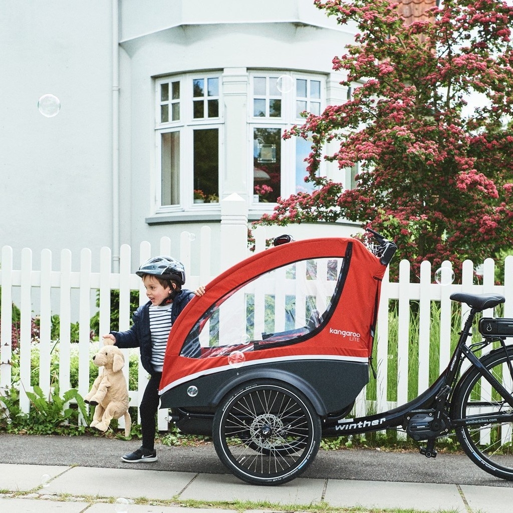 1:1 zugeschnittenes Bild - Winther Kangaroo Lite Lastenrad mit rotem Verdeck. Ein Kind mit Helm steht neben dem Fah